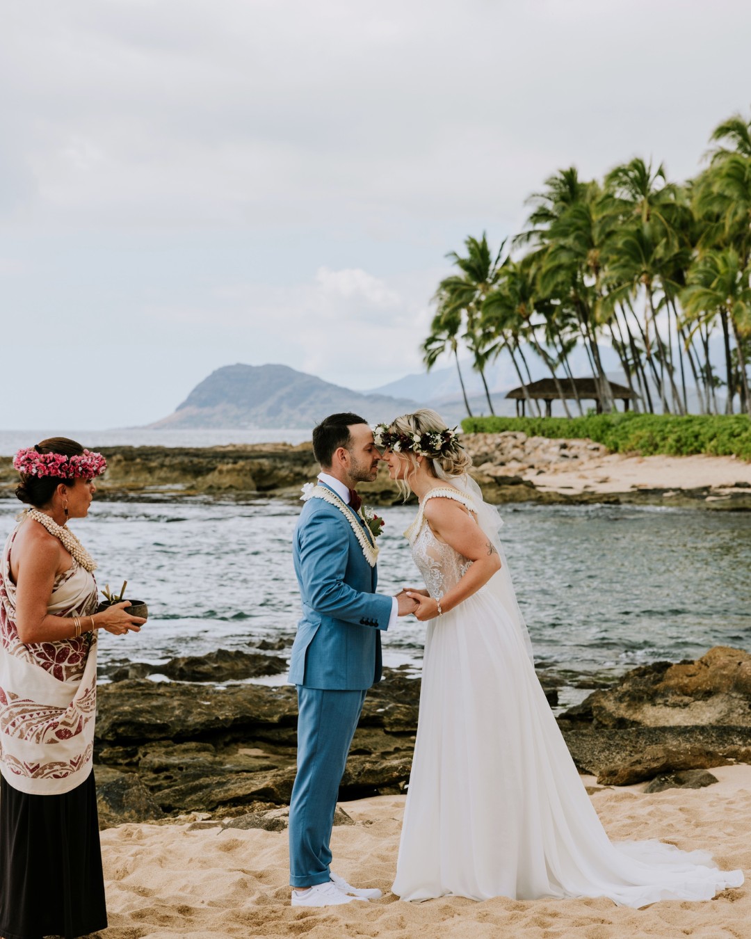 Destination Wedding Destination Wedding auf Hawaii mit einem Brautpaar bei einer traditionellen Zeremonie am Strand, begleitet von einer hawaiianischen Zeremonienleiterin vor tropischer Kulisse mit Palmen und Meer.