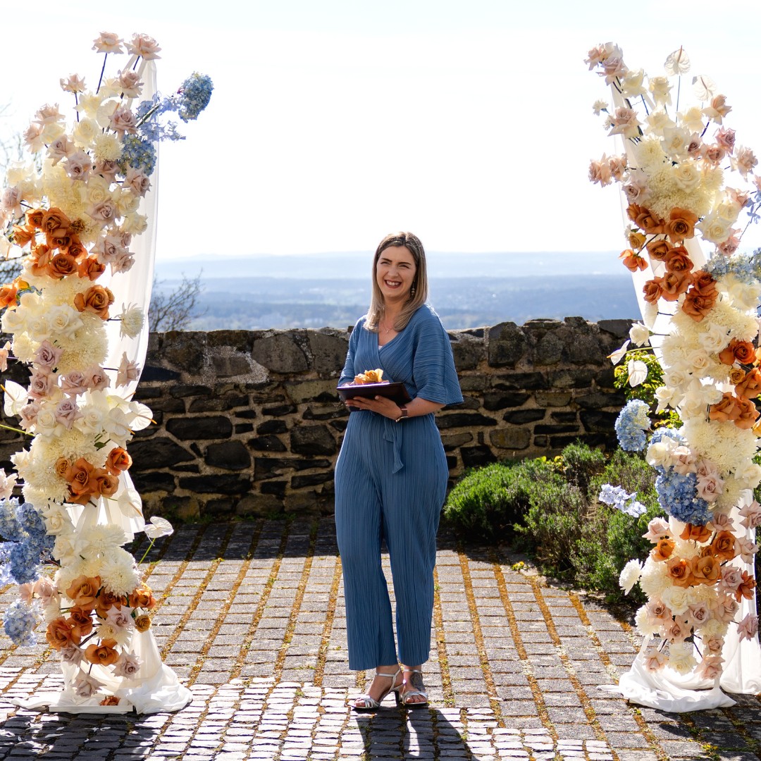 Janine Rhode Traurednerin freie Trauung Hamburg2 „Janine Rhode, Traurednerin für freie Trauung in Hamburg, steht lächelnd zwischen zwei floralen Traubögen vor einer Naturkulisse mit Blick ins Weite